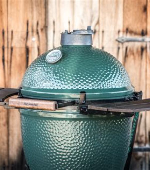 Grüner Big Green Egg Keramikgrill in großer Ausführung mit aufgeklappten Seitentischen vor Holzwand.