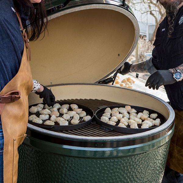Zwei Personen öffnen einen sehr großen Big-Green-Egg-Keramikgrill, in dem zwei gusseiserne Pfannen mit gewürzten Jakobsmuscheln liegen.