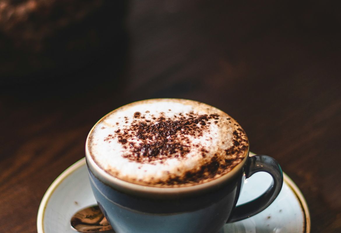 Cappuccino in blauer Tasse auf Holztisch mit Milchschaum und Schokostreuseln in gemütlicher Café-Atmosphäre.