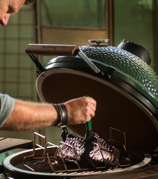 Person glasiert ein großes Stück Fleisch im offenen Big Green Egg Grill, Keramikdeckel geöffnet, intensives Grillambiente.