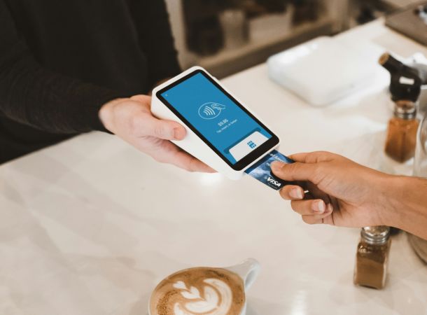 Gast bezahlt kontaktlos mit Karte an einem mobilen Terminal, im Vordergrund steht eine Tasse Cappuccino.