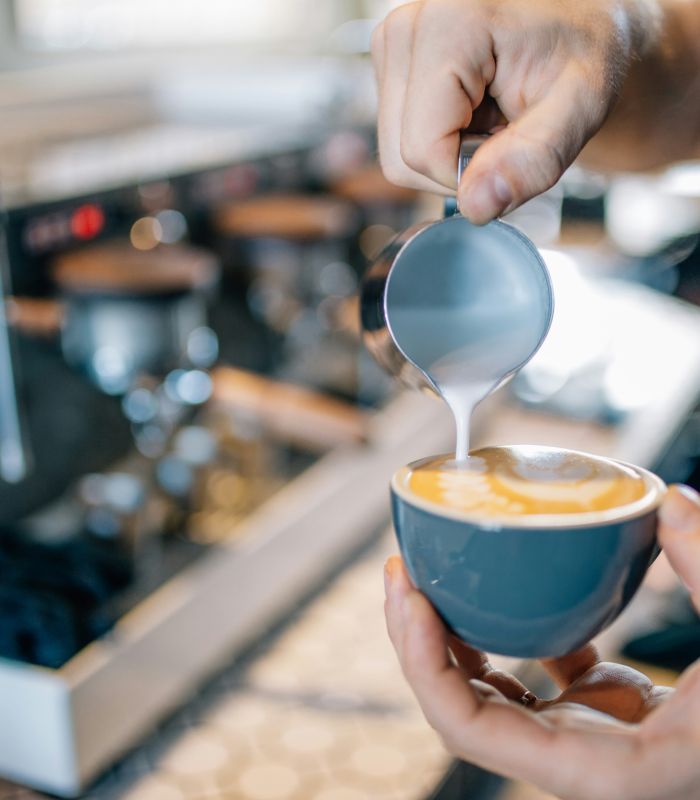Barista gießt cremigen Milchschaum aus einer kleinen Kanne in eine Cappuccino-Tasse vor einer Siebträgermaschine.