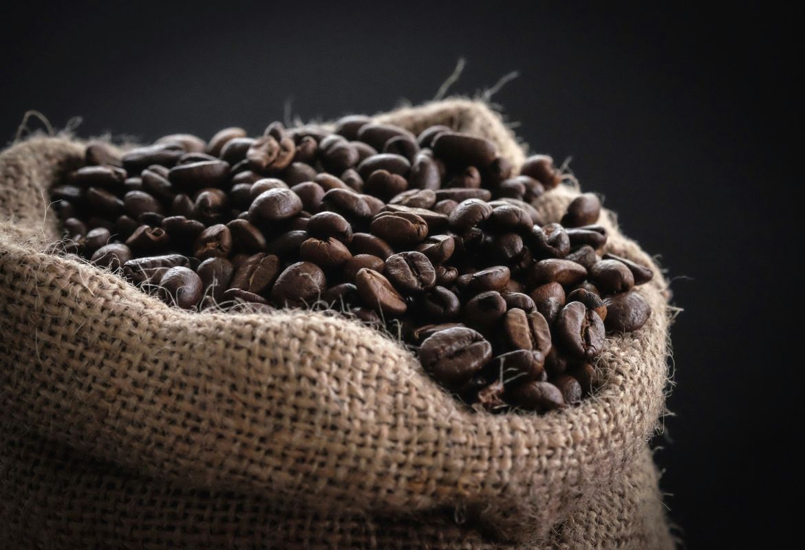 Offener Jutesack gefüllt mit dunkel gerösteten Kaffeebohnen vor dunklem Hintergrund.