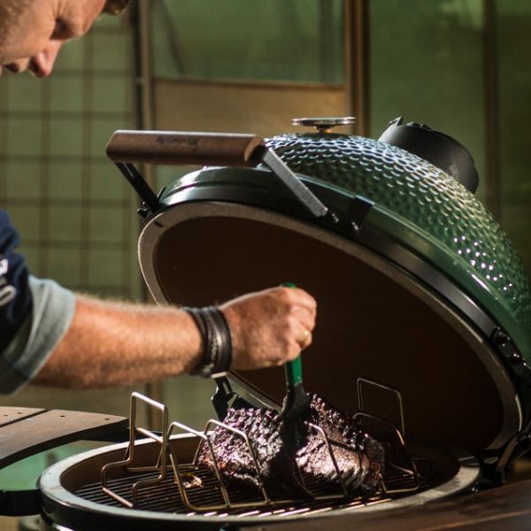 Person glasiert ein großes Stück Fleisch im offenen Big Green Egg Grill, Keramikdeckel geöffnet, intensives Grillambiente.
