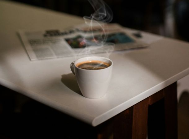 Heiße Tasse Kaffee mit aufsteigendem Dampf auf einem Tisch neben einer geöffneten Zeitung.