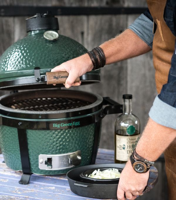 Grüner Big Green Egg Grill, geöffnet, während ein Mann mit Lederschürze Zutaten vorbereitet.