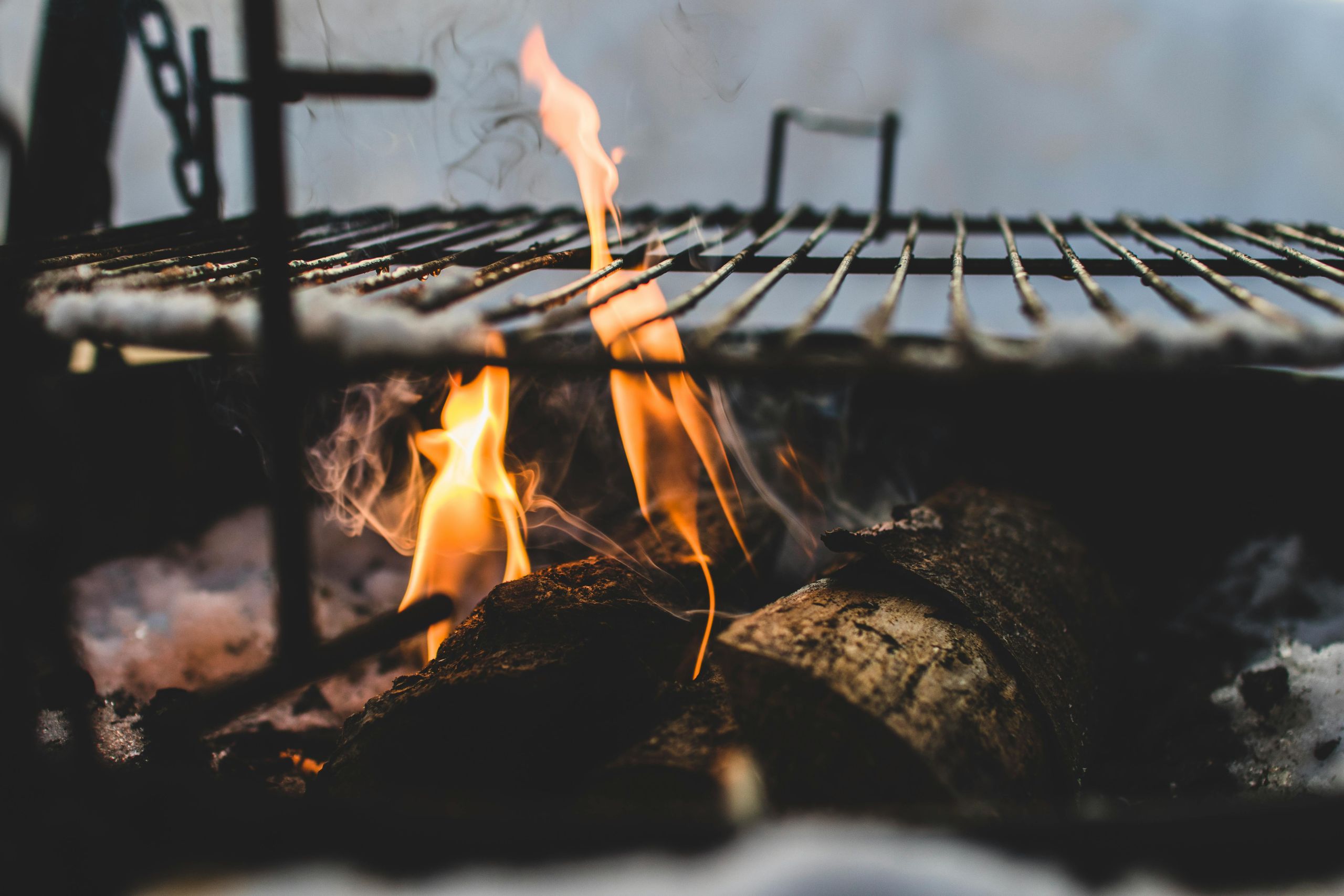 Brennendes Holz unter einem Grillrost mit offenen Flammen und aufsteigendem Rauch im Nahaufnahme-Detail.
