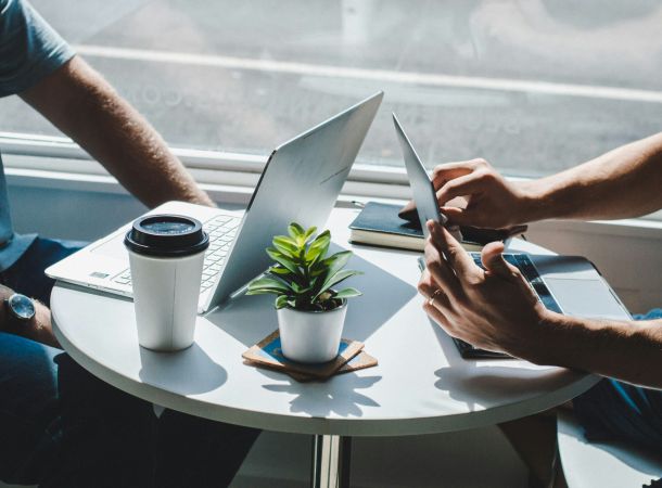 Zwei Personen arbeiten mit Laptops an einem kleinen Cafétisch, daneben steht ein Coffee-to-go-Becher und eine kleine Pflanze.