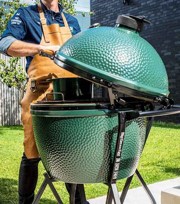 Großer grüner Keramikgrill Big Green Egg XLarge im Garten, geöffnet und von einer Person mit Lederschürze bedient.