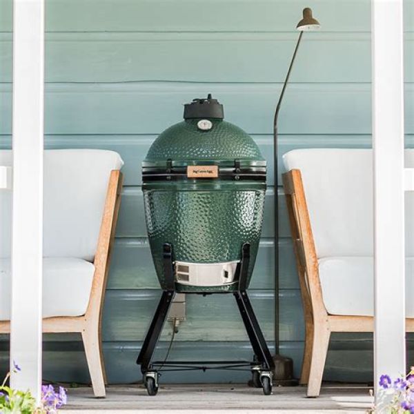Grüner Big-Green-Egg-Keramikgrill in mittlerer Größe auf Rollen, platziert zwischen zwei weißen Sesseln auf einer Veranda.