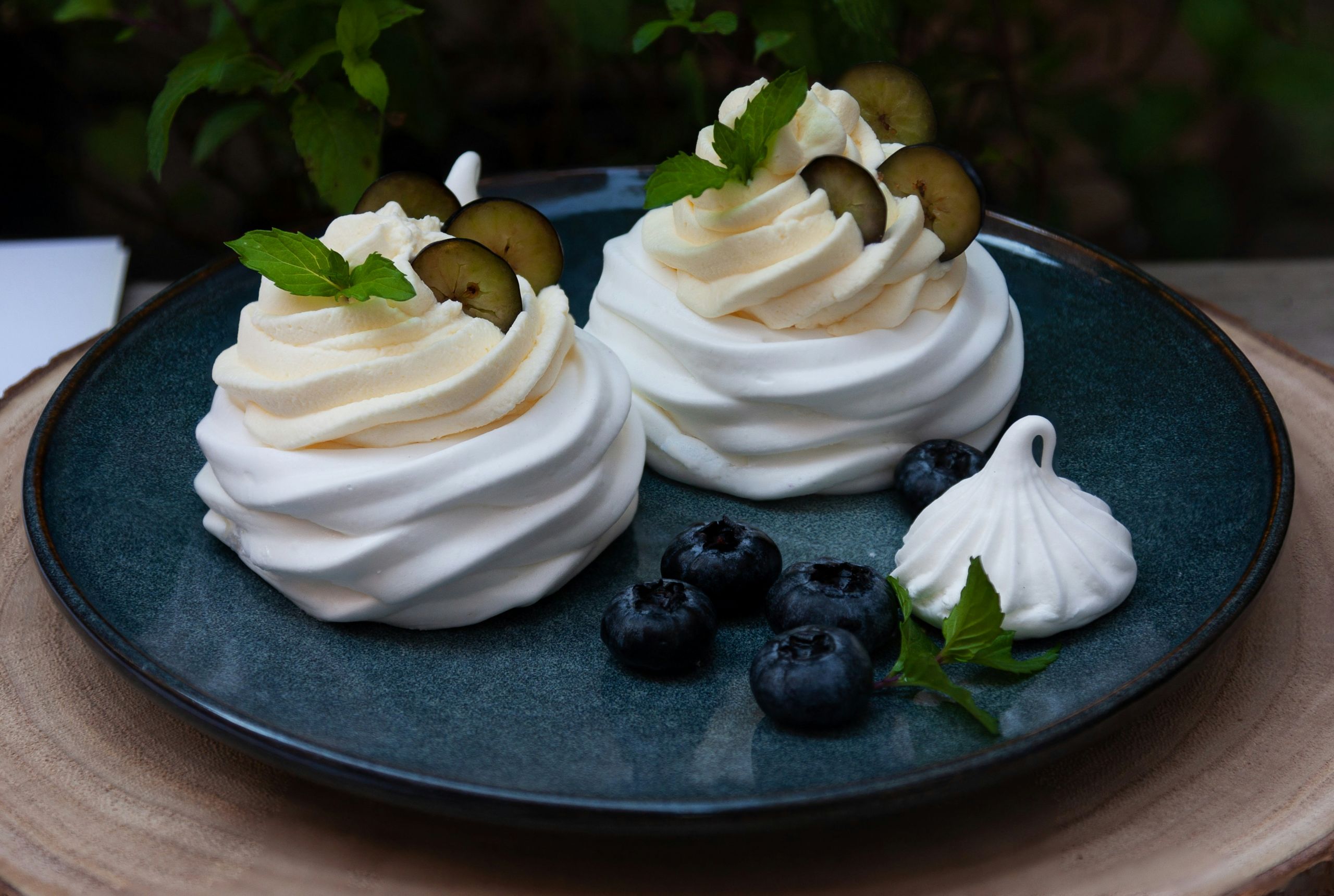 Zwei Pavlovas mit Sahne, Trauben und Minze auf dunklem Teller, garniert mit frischen Blaubeeren und kleinem Baiser.