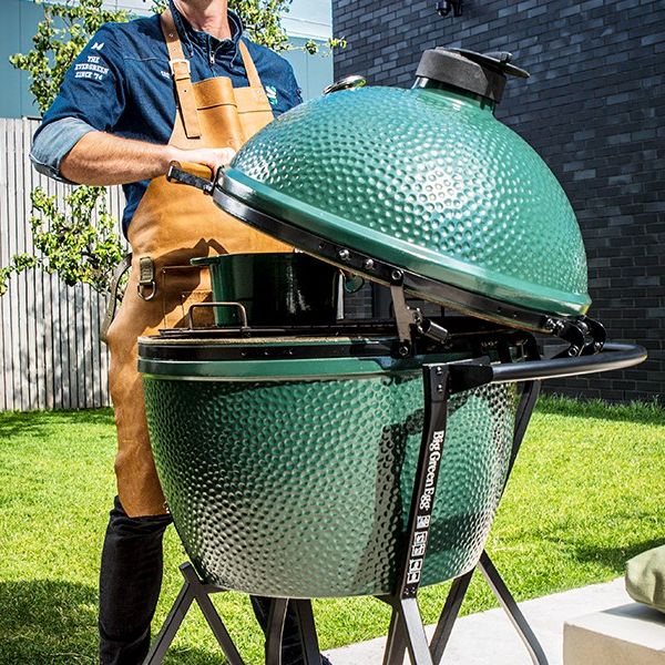 Großer grüner Keramikgrill Big Green Egg XLarge im Garten, geöffnet und von einer Person mit Lederschürze bedient.