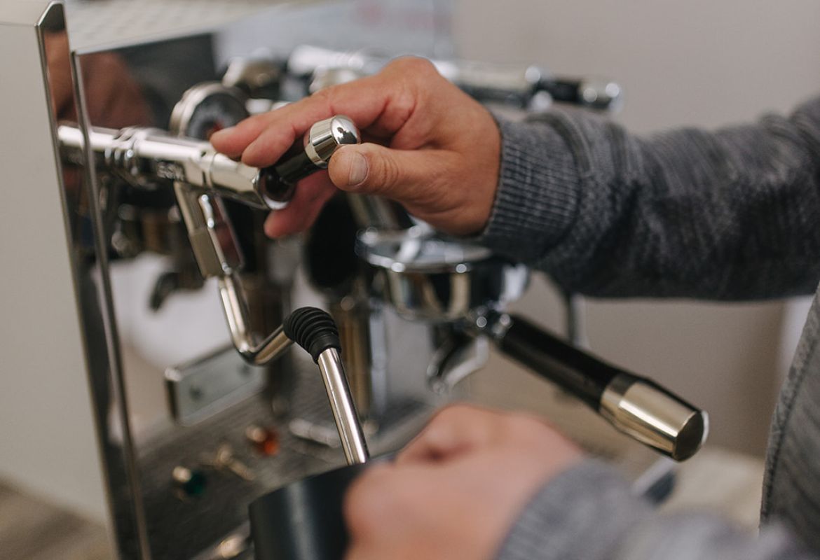 Barista schäumt Milch an der Espressomaschine auf und hält ein Milchkännchen unter die Dampflanze.