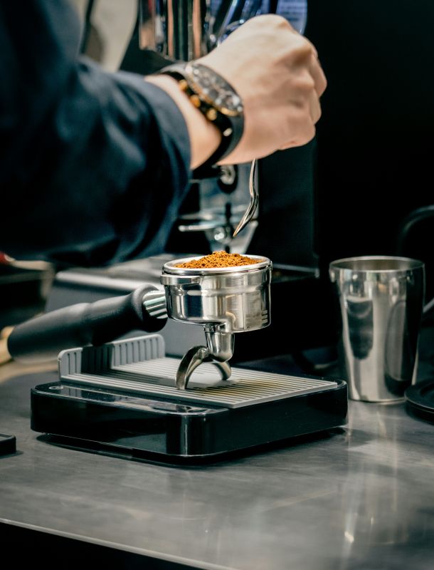 Barista füllt frisch gemahlenen Kaffee mit einem Löffel in einen Siebträger auf einer Tamperstation.