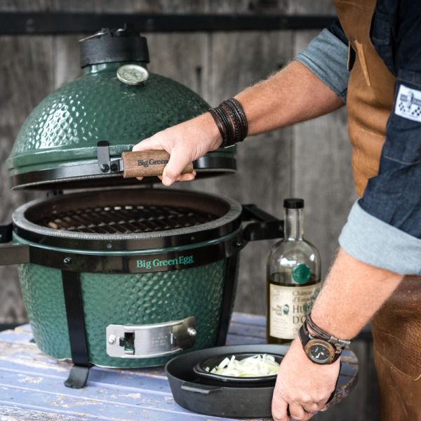 Grüner Big Green Egg Grill, geöffnet, während ein Mann mit Lederschürze Zutaten vorbereitet.