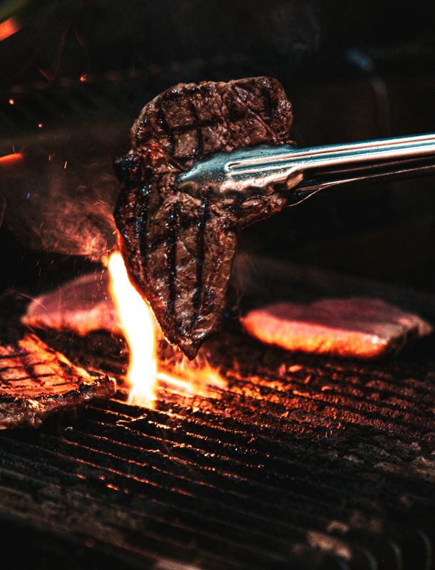 Steak wird mit Grillzange über offener Flamme auf einem heißen Grill gewendet, umgeben von Rauch und Glut.