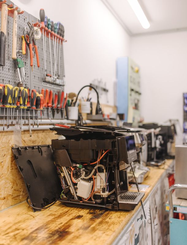 Geöffnete Kaffeemaschine zur Reparatur in der Werkstatt, umgeben von Werkzeugen an der Wand.