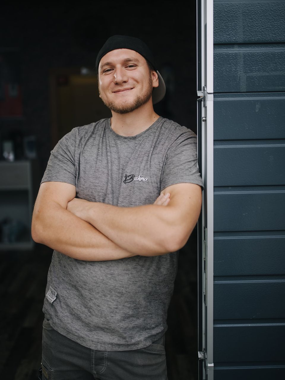 Junger Mann mit Basecap und verschränkten Armen lehnt lächelnd im Türrahmen einer Werkstatt oder Kaffee-Servicehalle.