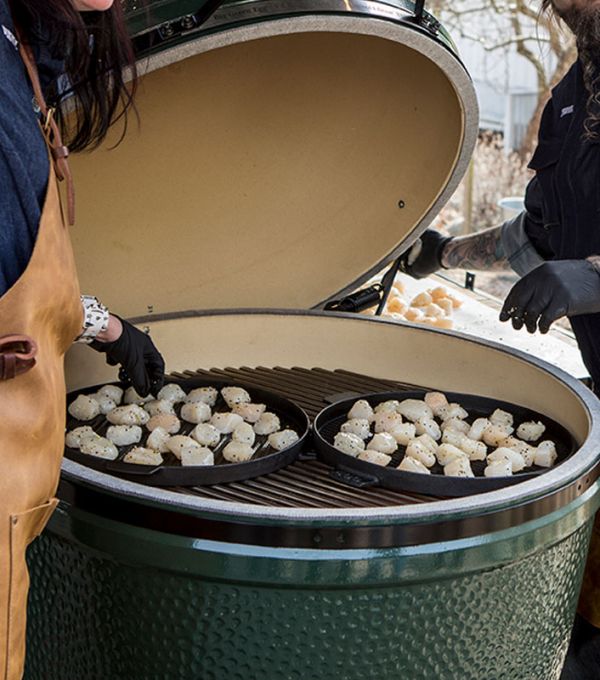 Zwei Personen öffnen einen sehr großen Big-Green-Egg-Keramikgrill, in dem zwei gusseiserne Pfannen mit gewürzten Jakobsmuscheln liegen.
