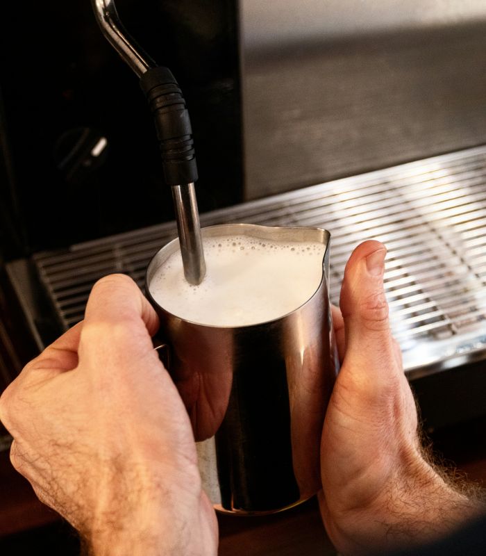 Barista schäumt Milch mit Dampflanze in einem Edelstahlkännchen auf einer Espressomaschine auf.