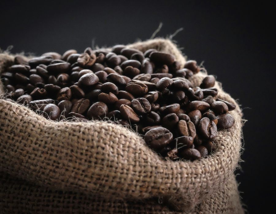 Offener Jutesack gefüllt mit dunkel gerösteten Kaffeebohnen vor dunklem Hintergrund.