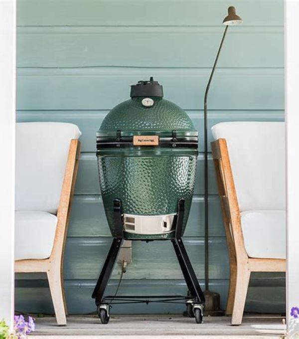 Grüner Big-Green-Egg-Keramikgrill in mittlerer Größe auf Rollen, platziert zwischen zwei weißen Sesseln auf einer Veranda.